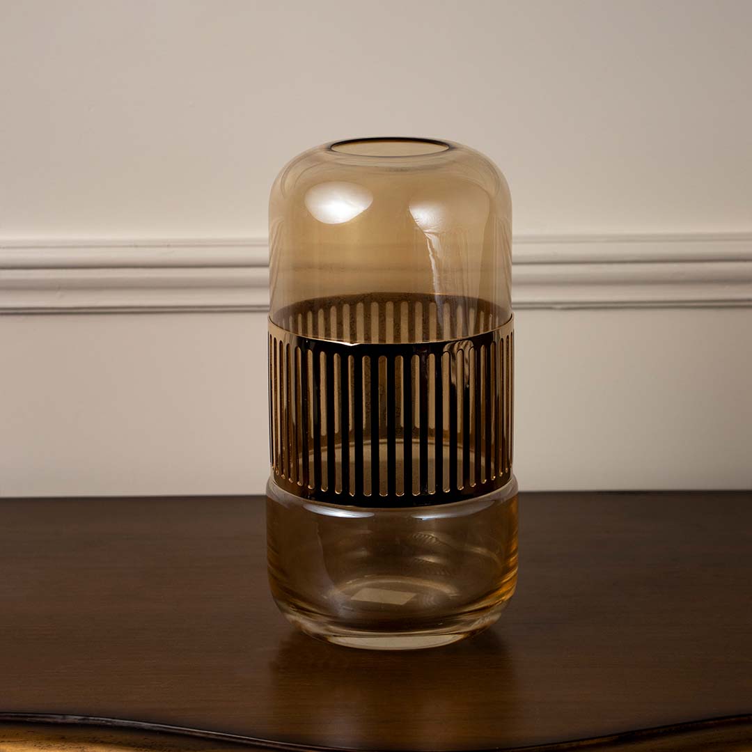 Amber glass decorative jar with vertical metal accents placed on wooden table against neutral wall background creating warm ambient decor lighting.