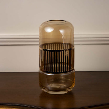 Amber glass decorative jar with vertical metal accents placed on wooden table against neutral wall background creating warm ambient decor lighting.