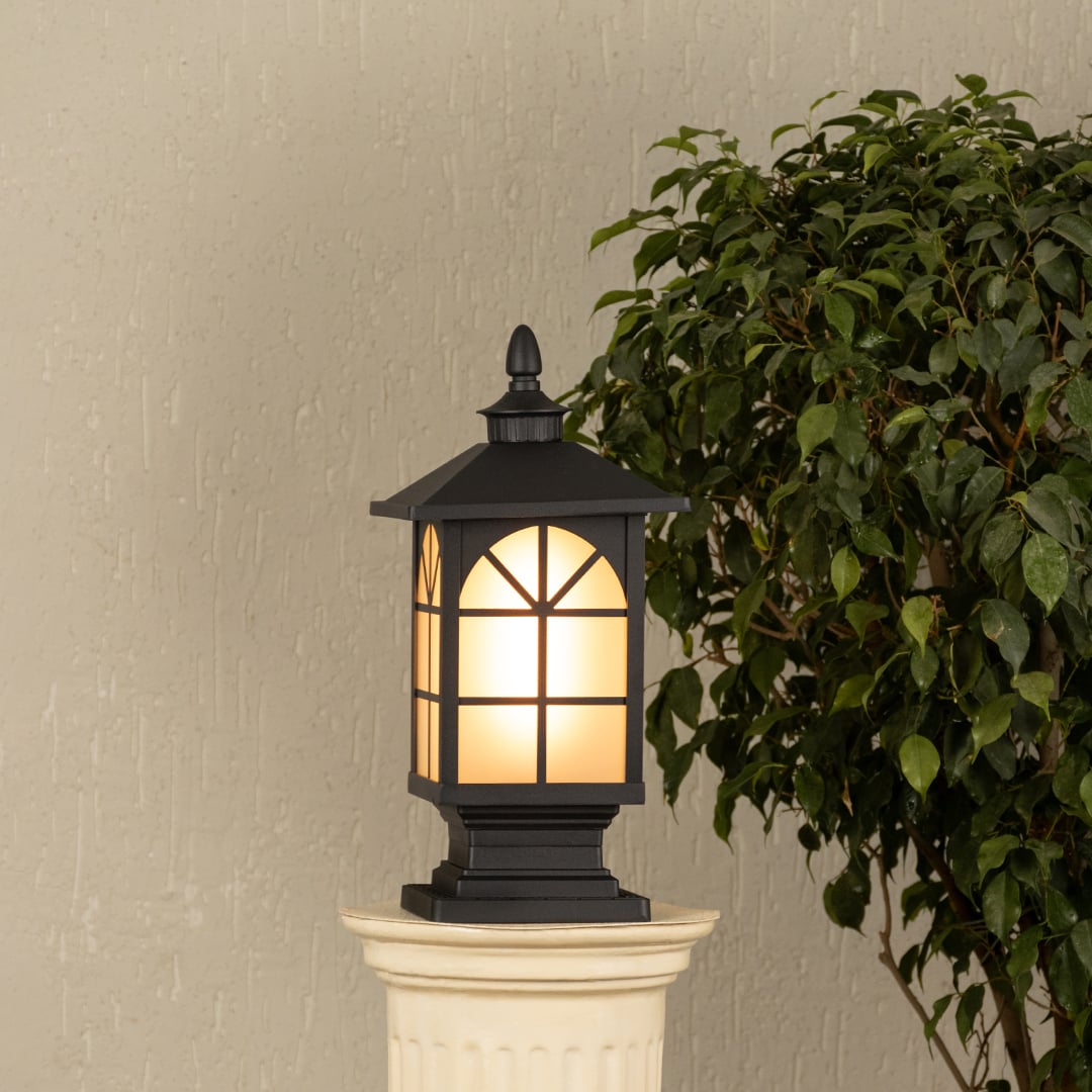 Black outdoor gate light with warm glowing panels styled beside plants for elegant exterior decor ambiance.