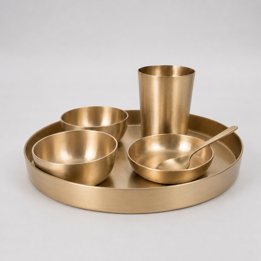 A set of matte brass tableware including a round tray, a tumbler, two bowls, and a spoon, arranged on a light gray background.