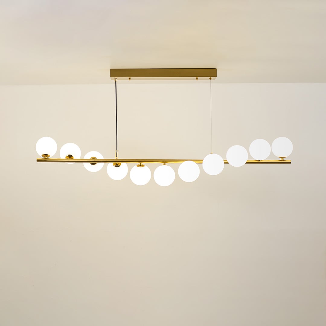 Gold linear chandelier with staggered white globes in a front view against a white wall background with lights on perfect for designer lighting décor.