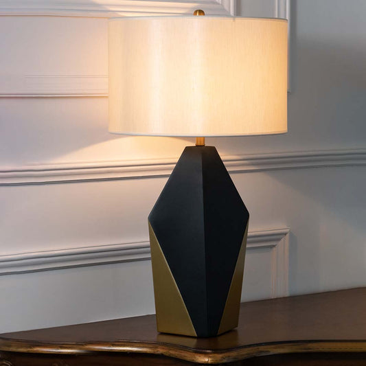 Black geometric table lamp with fabric drum shade and gold accents creating sophisticated modern ambient lighting indoors.