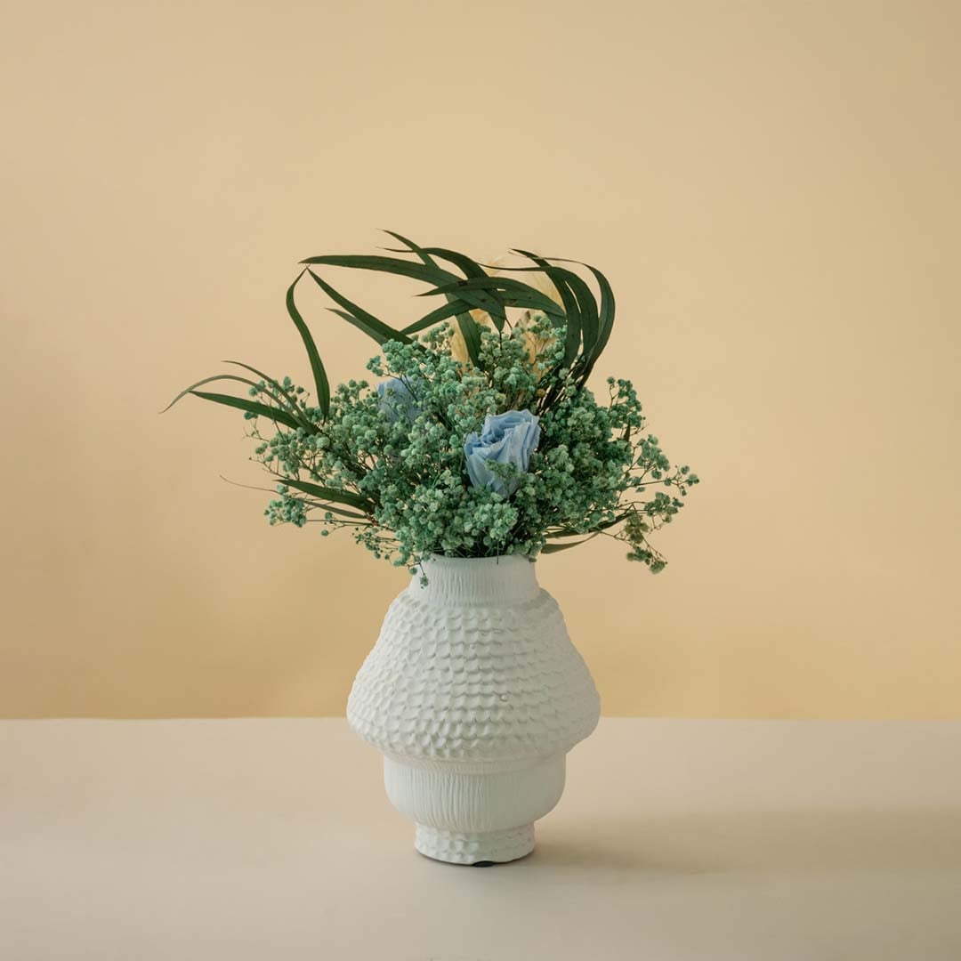 Textured white ceramic vase styled with green and blue artificial flowers placed on a wooden table beside a cup and candle for cozy home decor.