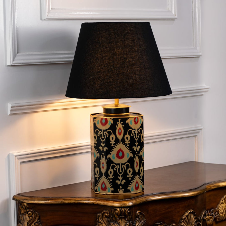 Table lamp with black fabric shade and decorative black base featuring gold and red traditional pattern placed on wooden side table.