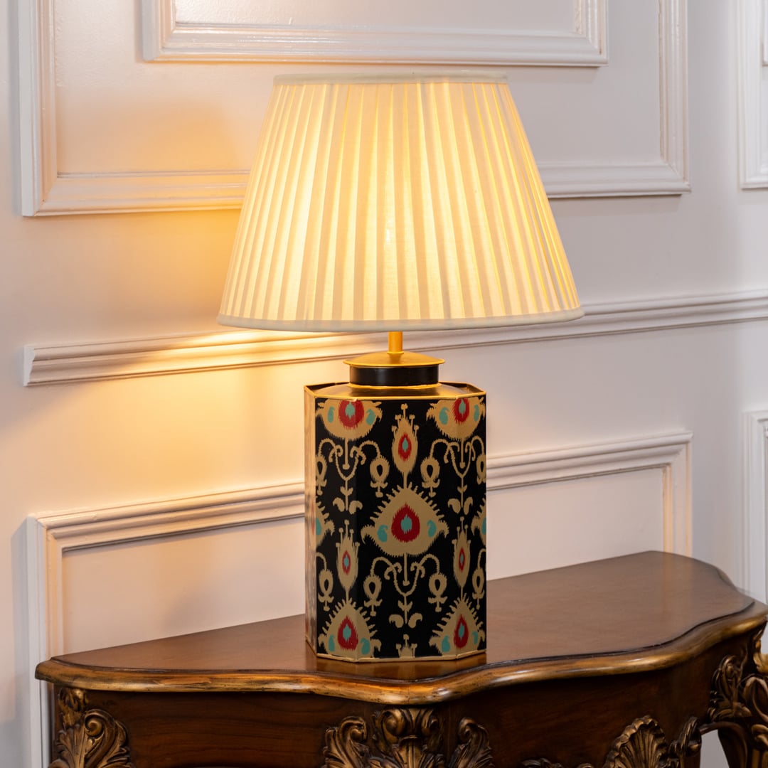 Elegant table lamp with pleated white shade and black ceramic base featuring intricate gold and red ethnic patterns on a wooden table.