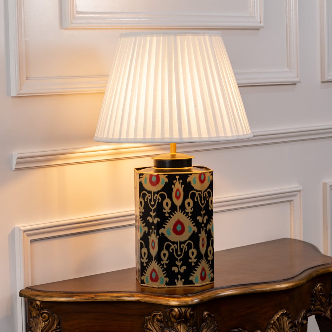 Table lamp with white pleated shade and black ceramic base featuring gold and folk inspired pattern placed on classic wooden table.