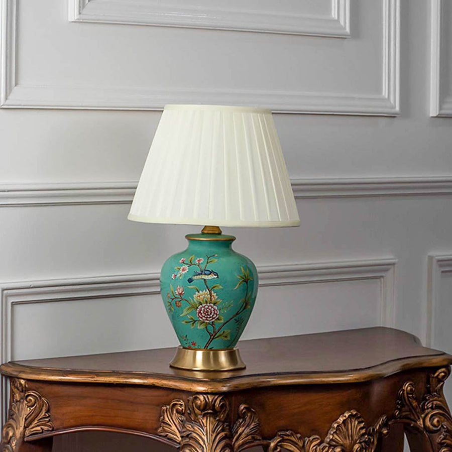 Elegant green ceramic table lamp turned on with golden base floral detailing and pleated shade on white background decor.