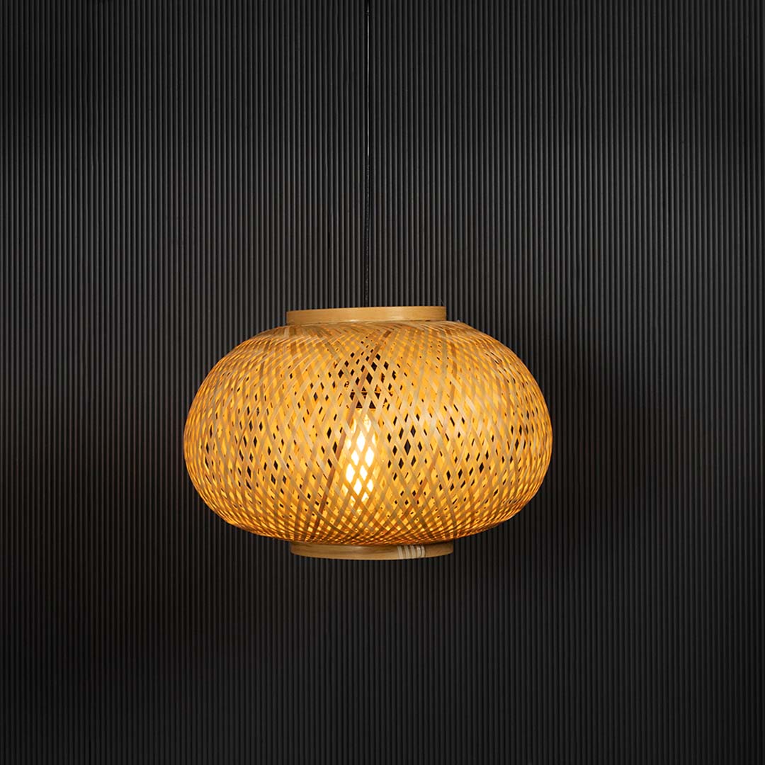 Round rattan pendant light with woven bamboo shade turned on glowing warmly against black background.