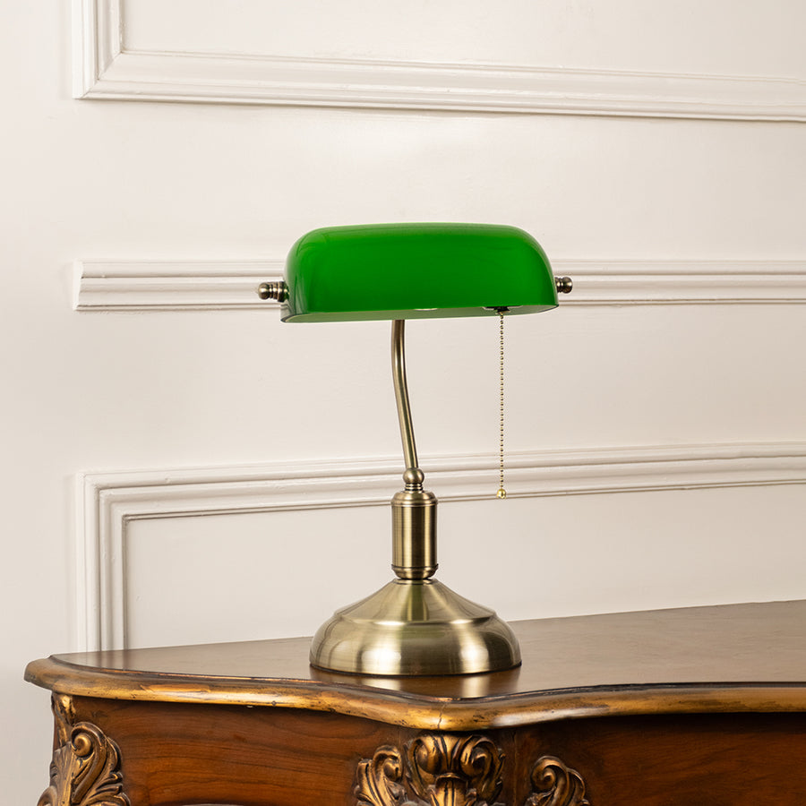 Green banker table lamp with brass metal base displayed on a wooden console table offering classic desk lighting.