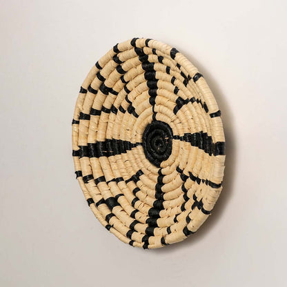 A side angle view of the handwoven natural and black rattan wall decor plate showing its raised rim and detailed weave for rustic artisan inspired home styling.