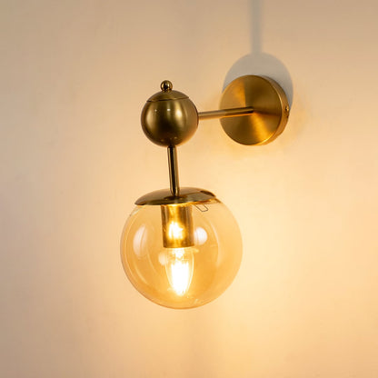 Brushed gold wall light with amber glass globe glowing on white background adding warm elegant lighting to modern living bedroom and hallway interiors.
