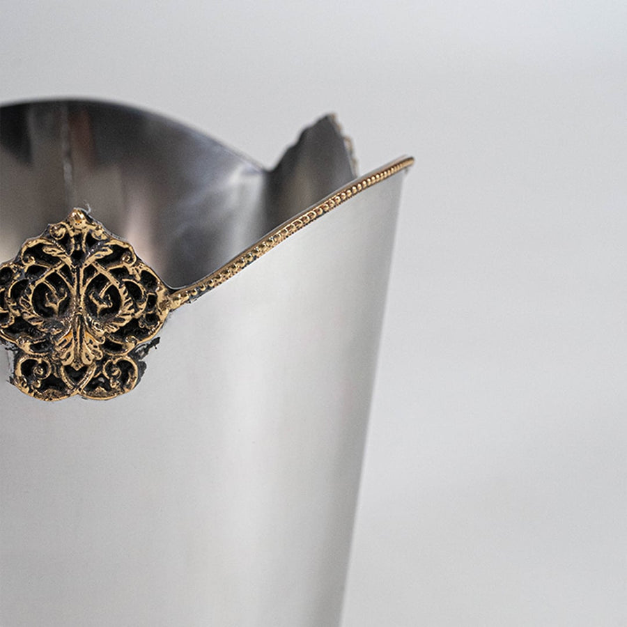 Silver metal waste bin with gold detailing placed beside wooden table and curtain on wooden floor for home decor.