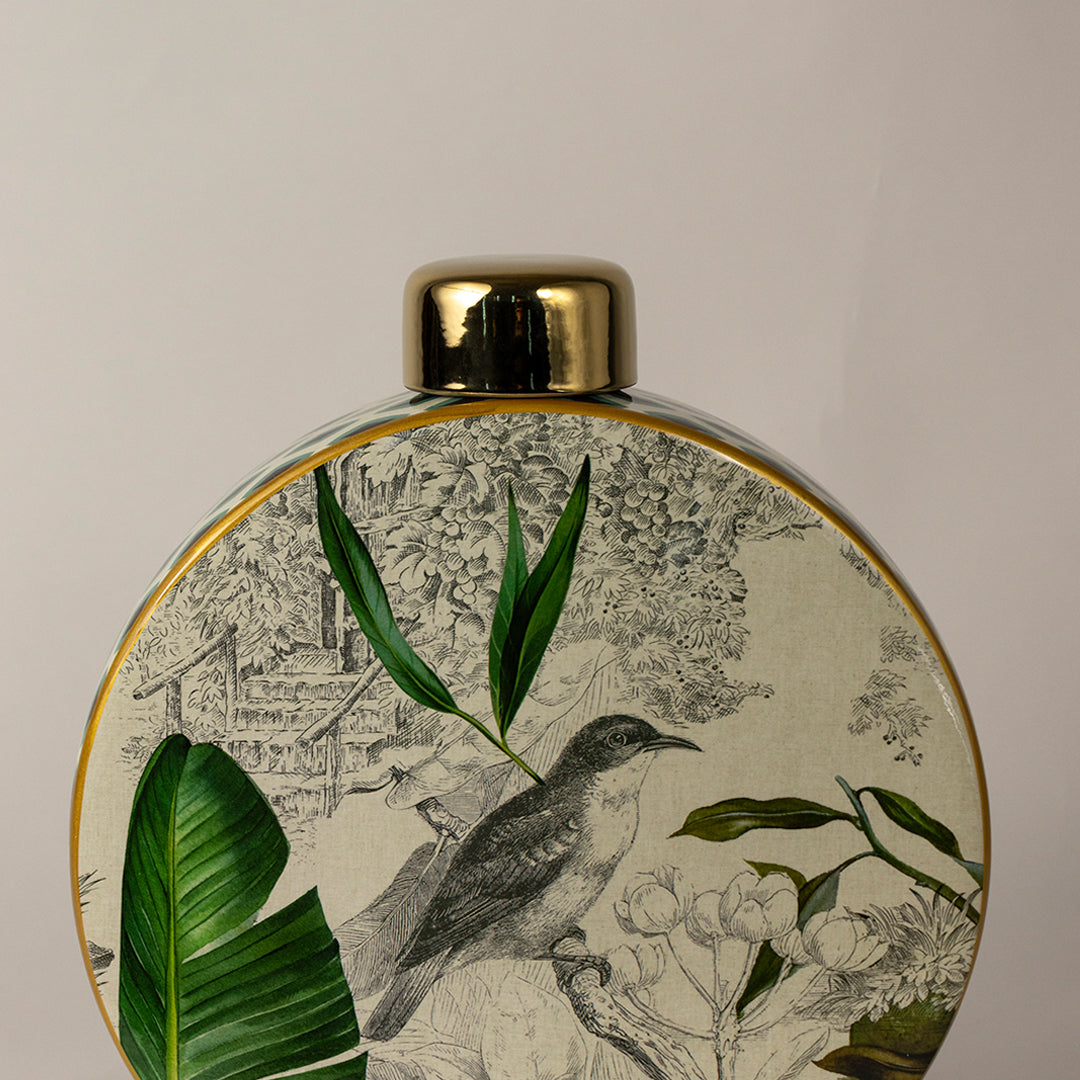 Round ceramic vase with green botanical leaf artwork and gold rim placed on white surface against neutral background closeup showcasing detailed nature inspired decor design.