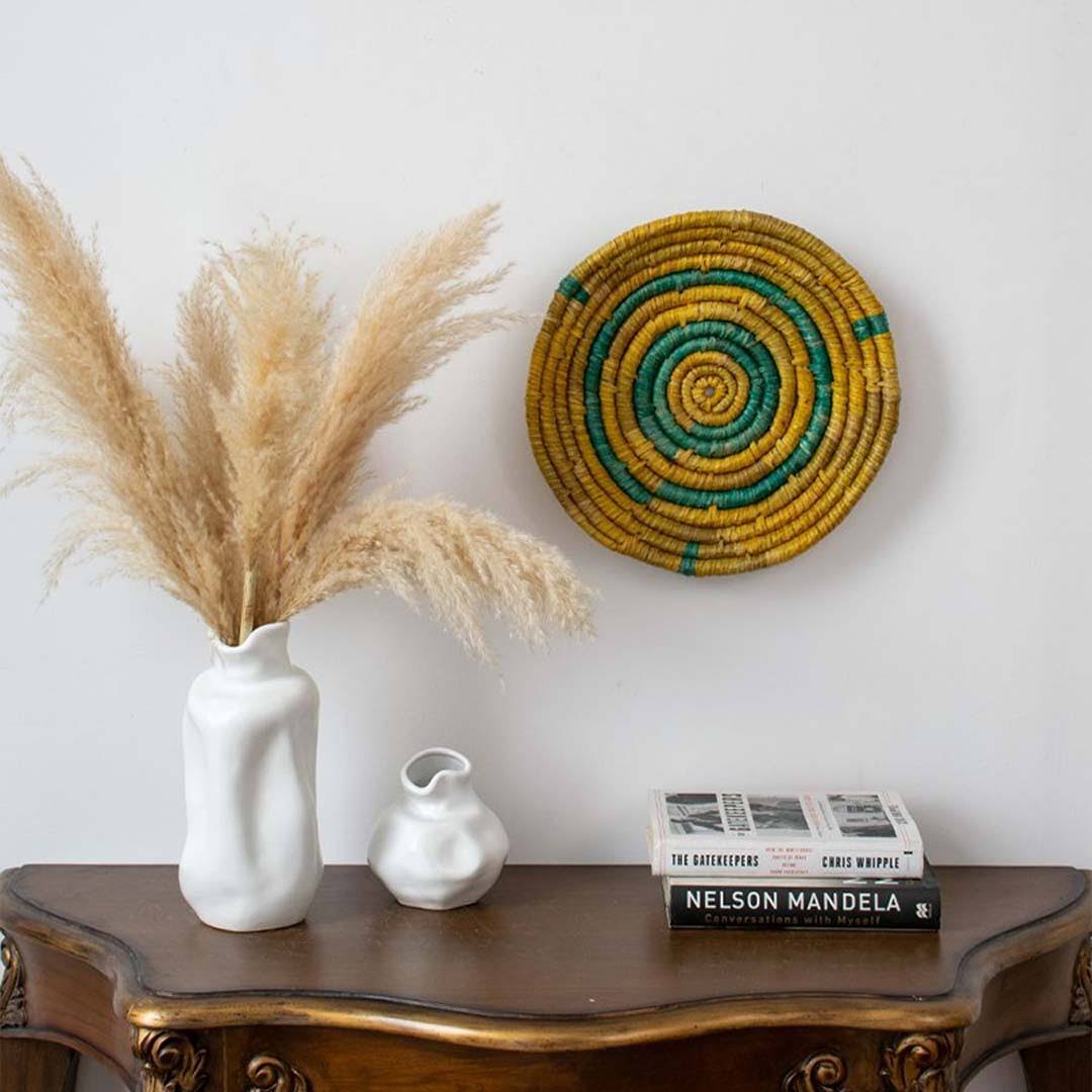 Decorative setup with pampas grass in a white vase, small white vase, books, and a woven wall hanging on a wooden table.
