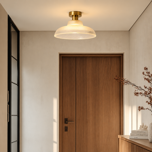 A modern ceiling light fixture with a ribbed frosted glass dome and a brushed brass ceiling mount, shown illuminated in a minimalist hallway with a wooden door and beige walls.