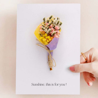 Hand holding a white greeting card with a mini yellow pink and purple dried flower bouquet isolated on a white backdrop.