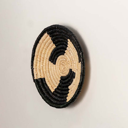 A side angle view of the natural and black rattan wall decor plate showing its raised rim and woven texture for warm artisan inspired wall styling.