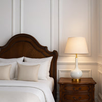 White ceramic table lamp placed on a wooden side table beside the bed illuminating the bedroom with warm light for cozy and luxurious home decor styling.