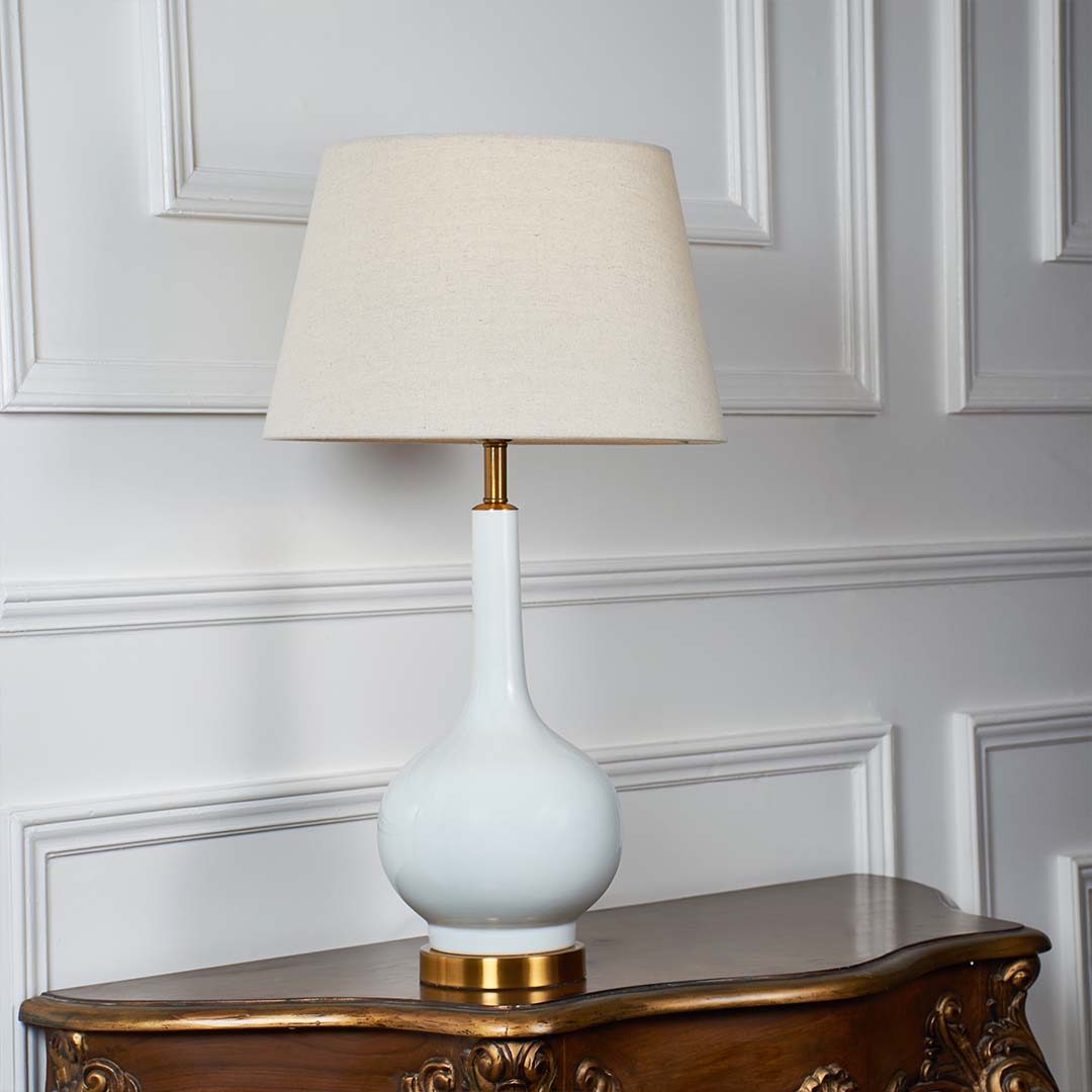 White ceramic table lamp with golden base and beige lampshade glowing warmly against a white wall background showcasing modern minimal design.