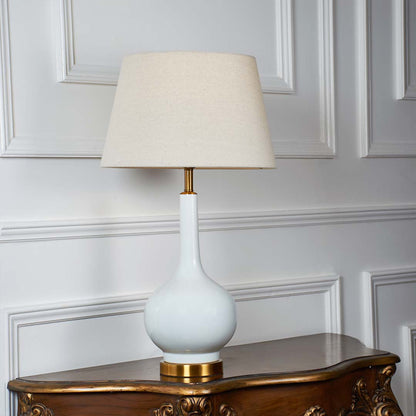 White ceramic table lamp with golden base and beige lampshade glowing warmly against a white wall background showcasing modern minimal design.