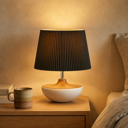 A modern table lamp with a pleated black shade, a light wood base, and a white ceramic bowl base, glowing on a wooden nightstand next to a ceramic mug.