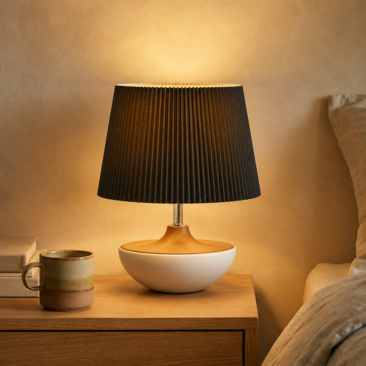 A modern table lamp with a pleated black shade, a light wood base, and a white ceramic bowl base, glowing on a wooden nightstand next to a ceramic mug.