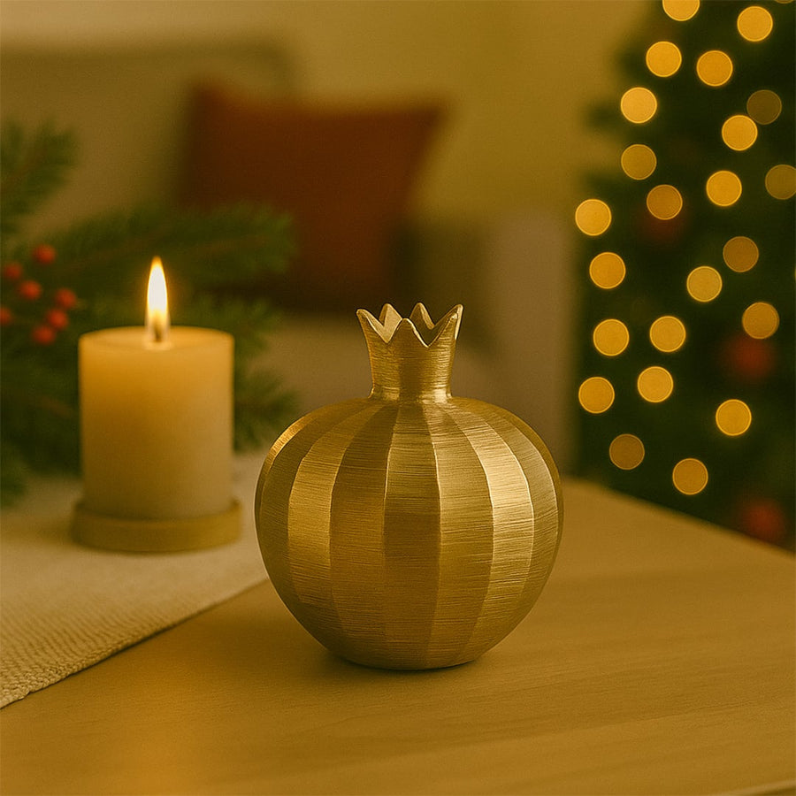 Warm festive setting showcasing a golden pomegranate decor placed on a wooden table with a glowing candle and softly lit Christmas tree in the background.