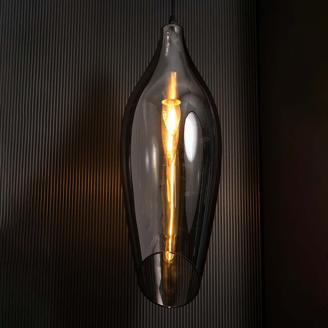 A smoked black sculpted glass pendant light with lights on casting a warm glow on a dark backdrop showcasing its tall curved silhouette.