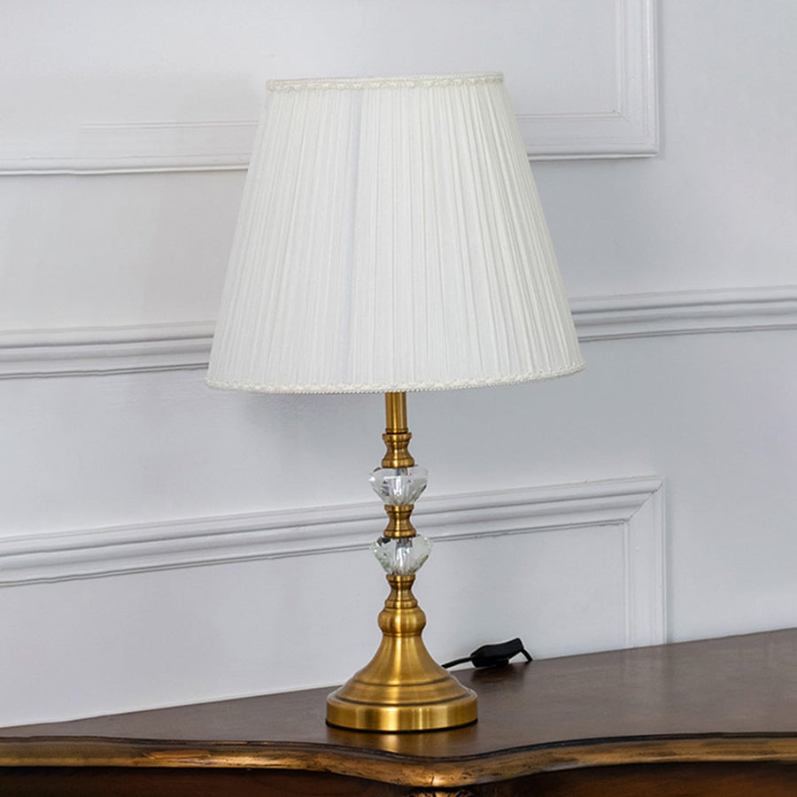 Crystal table lamp with gold accents and pleated white shade styled in elegant living room decor with light turned on for warmth.