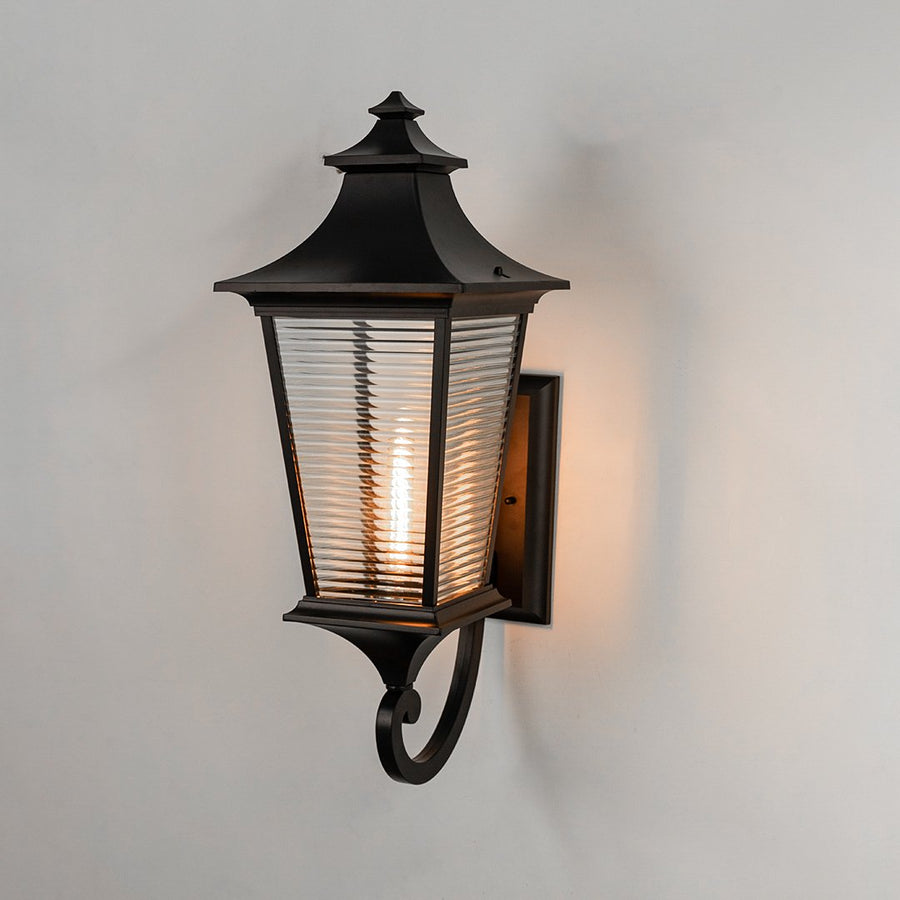 Black metal outdoor wall light on white background with horizontal ribbed glass shade illuminating warm patio decor.
