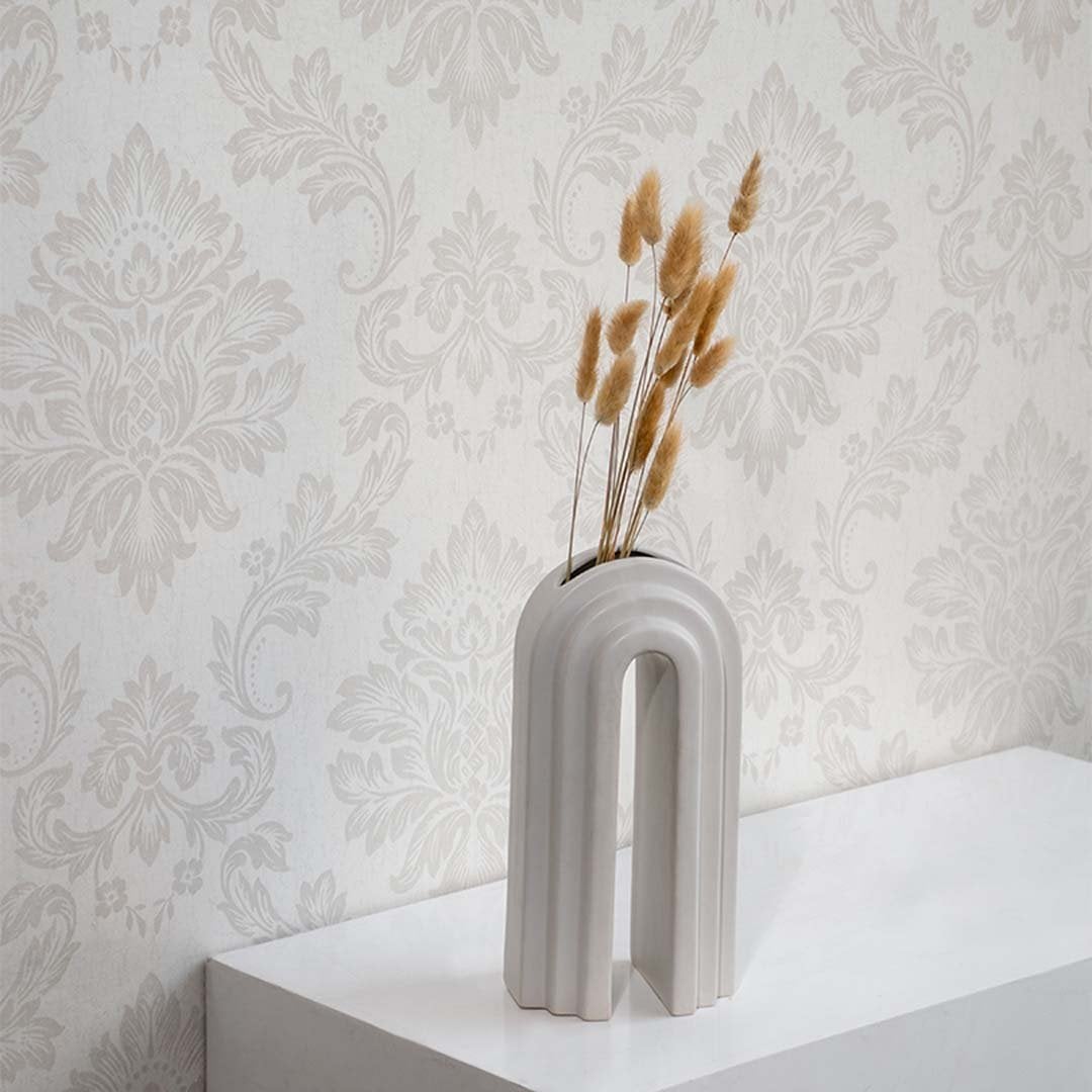 A modern arched ceramic vase holding dried grass stems displayed on a white surface against a patterned wallpaper backdrop creating elegant minimal botanical décor for interiors.