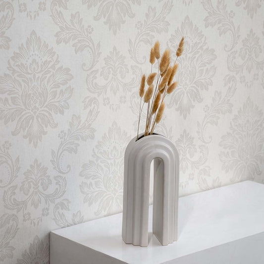 A modern arched ceramic vase holding dried grass stems displayed on a white surface against a patterned wallpaper backdrop creating elegant minimal botanical décor for interiors.