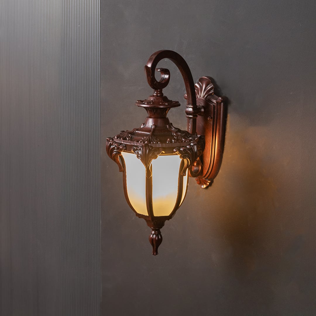 Outdoor wall lantern in bronze metal with frosted glass shade turned on glowing softly against plain black background.