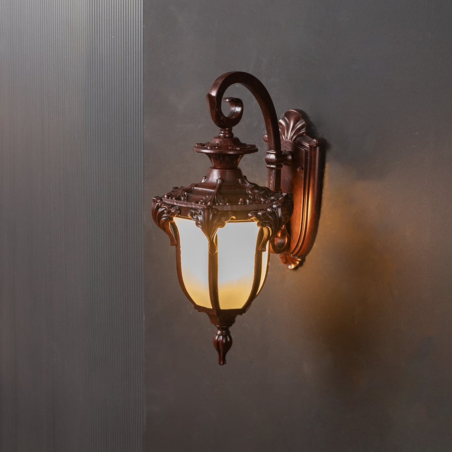 Outdoor wall lantern in bronze metal with frosted glass shade turned on glowing softly against plain black background.