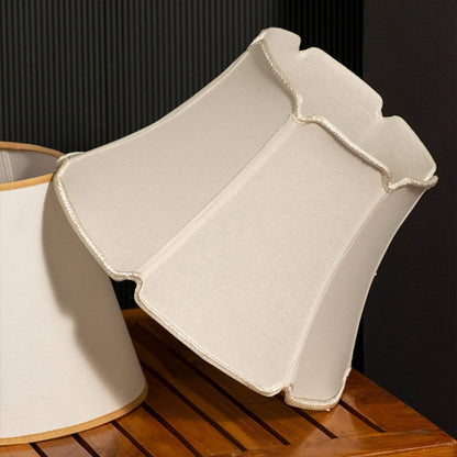 A soft beige fabric lampshade with a scalloped edge design displayed on a wooden surface with a black background creating a warm side view look.
