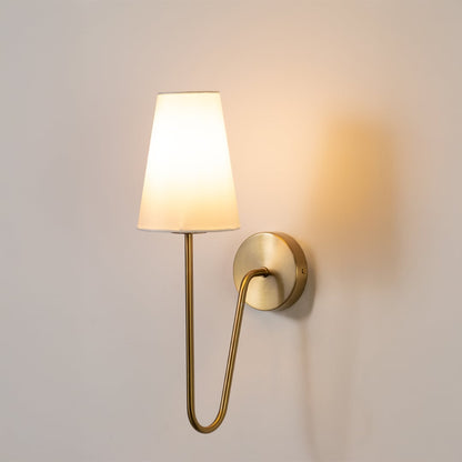 Gold metal wall light with a tall white fabric shade glowing softly on a clean white background for modern decor.