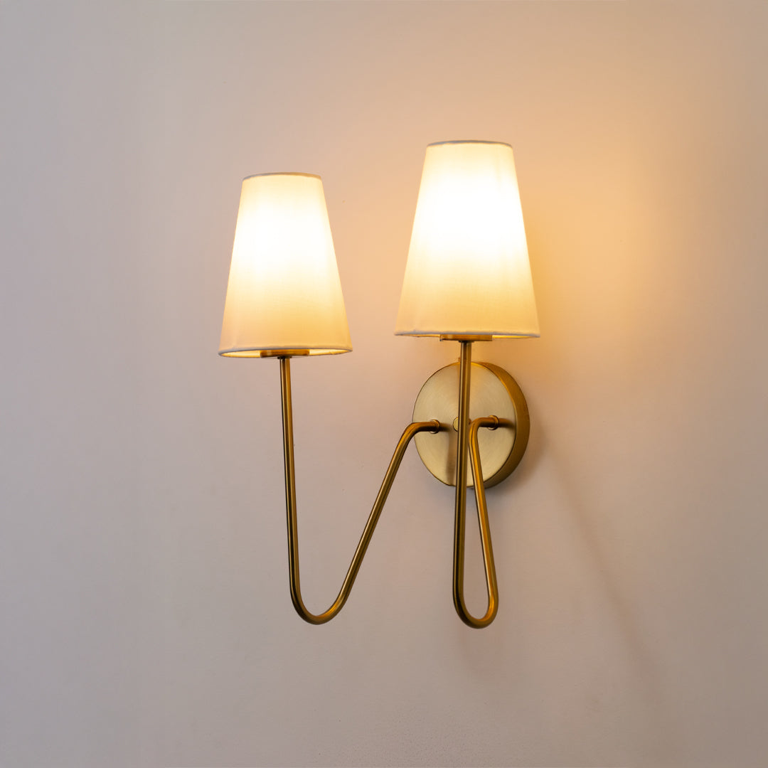 Closeup wall light in gold metal finish with dual fabric shades for bedroom or living room ambient lighting decor white background.