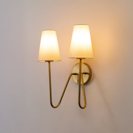 Closeup wall light in gold metal finish with dual fabric shades for bedroom or living room ambient lighting decor white background.