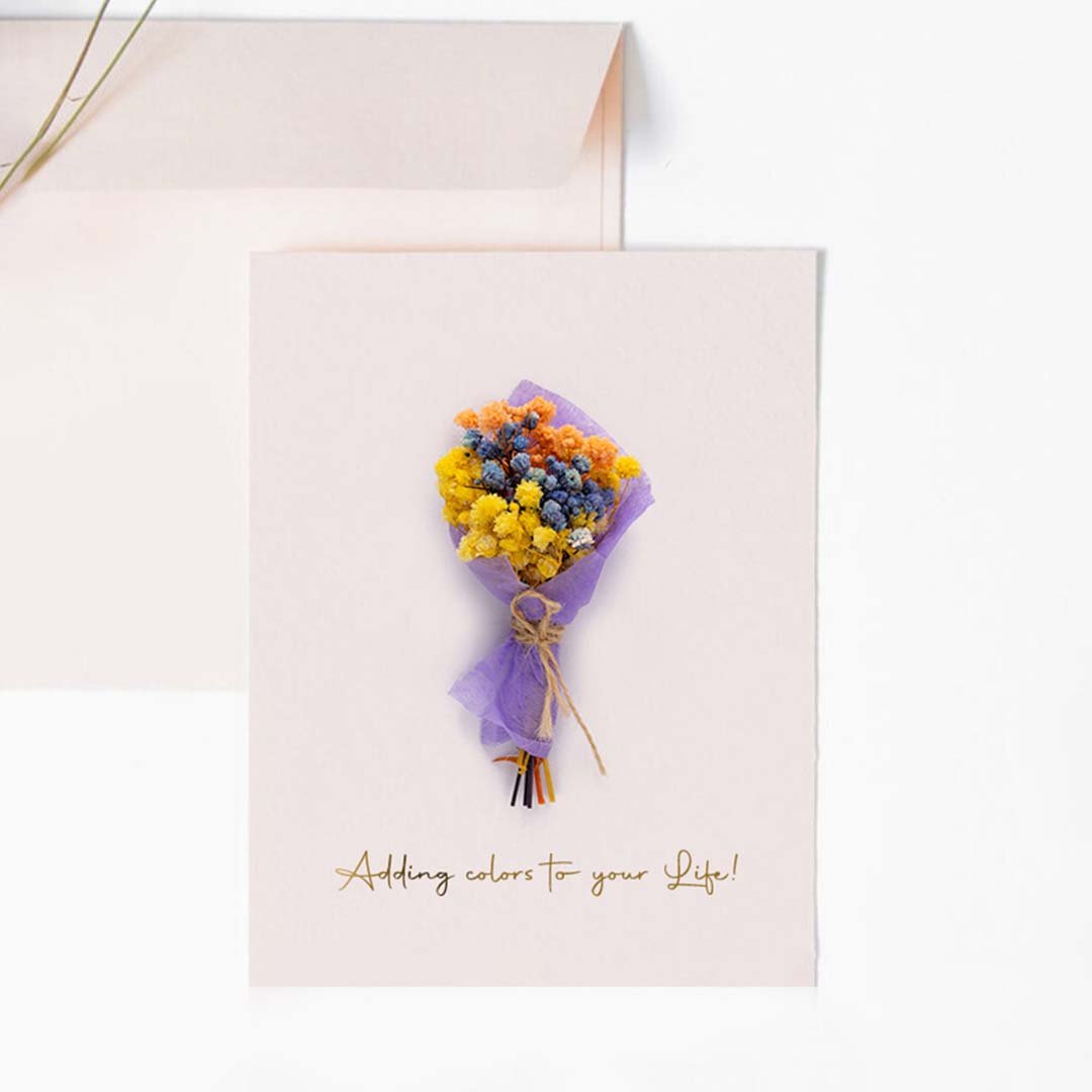 A handmade pink greeting card featuring a mini purple and yellow dried flower bouquet next to a matching light pink envelope.