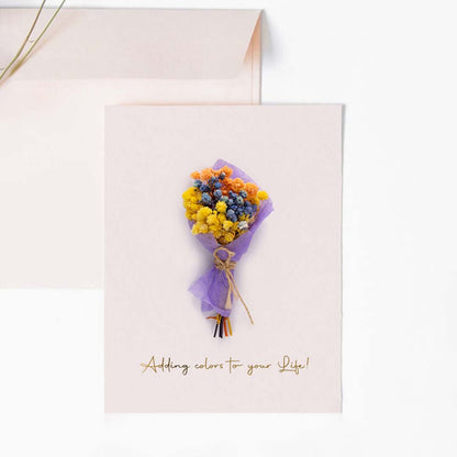A handmade pink greeting card featuring a mini purple and yellow dried flower bouquet next to a matching light pink envelope.