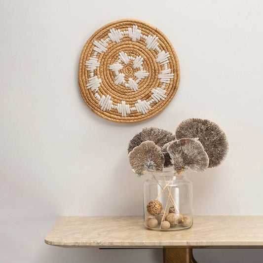 Natural seagrass wall plate with white geometric design arranged on a wall above a marble table with dried botanicals for styling.