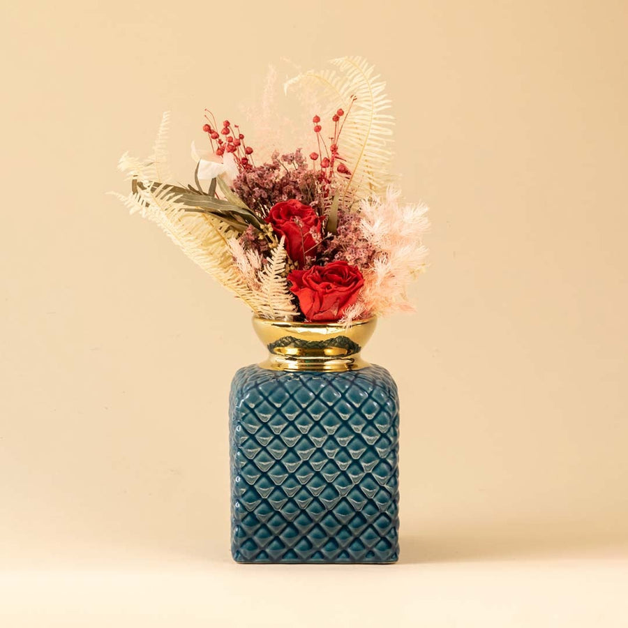 A blue textured ceramic vase with a gold top filled with red flowers displayed in a warm beige setting creating an elegant lifestyle decor look with soft natural light.