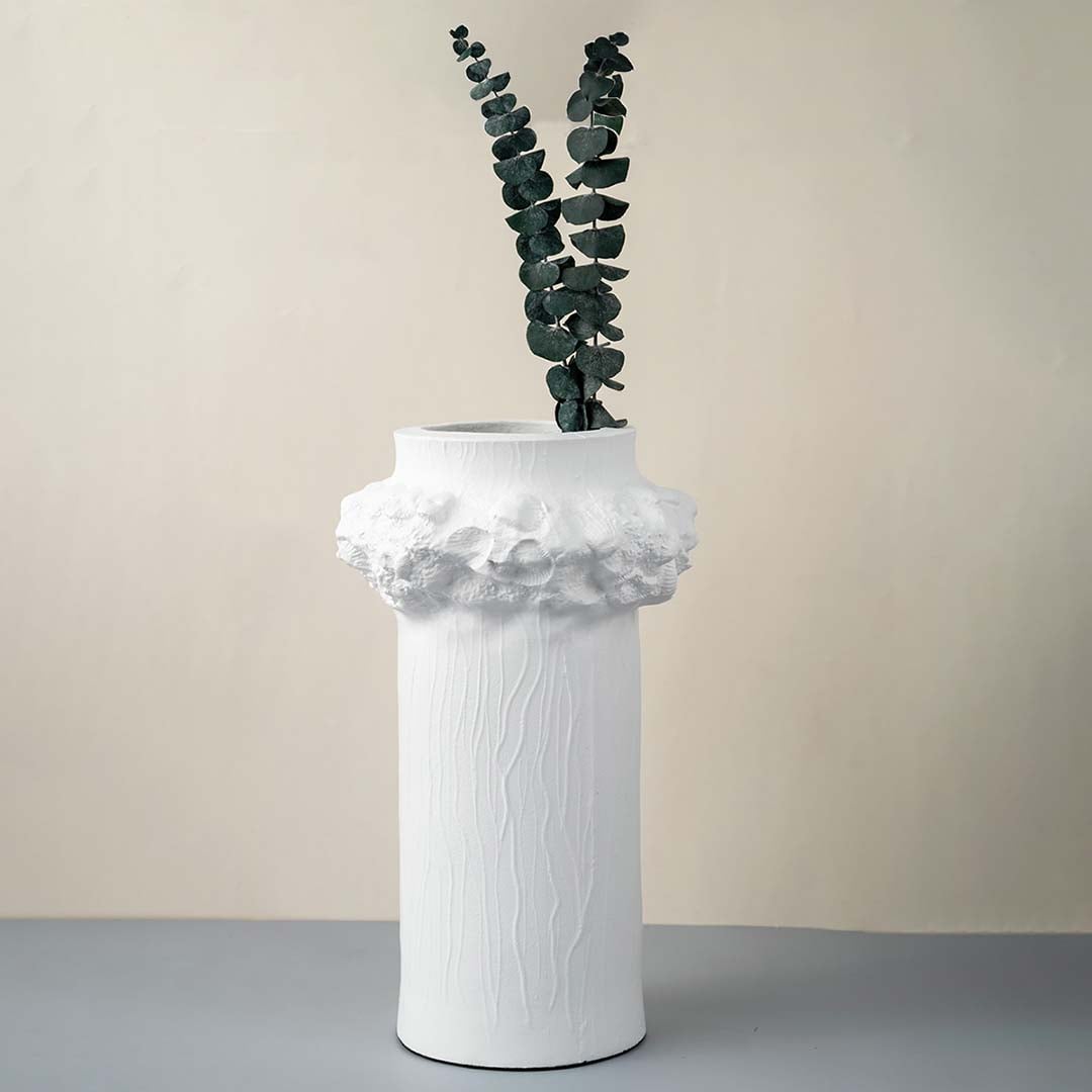 Tall white ceramic vase with textured surface and floral crown detail styled with green leaves for home decor.