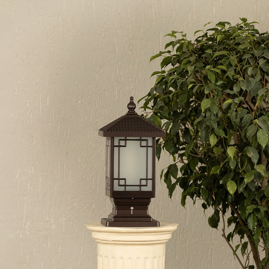 Black outdoor pillar light with ribbed glass Light On placed next to a lush green plant Lifestyle image for modern patio decor.