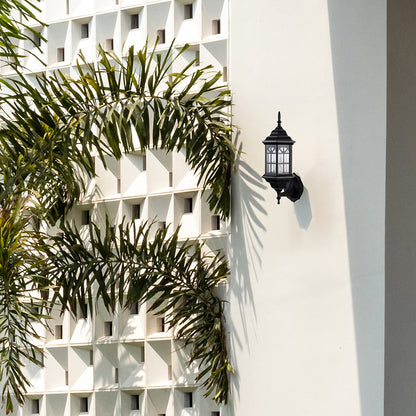 Black metal outdoor wall light lifestyle image with paneled glass shade mounted on white villa wall with palm trees.