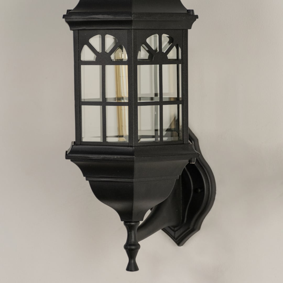 Black metal outdoor wall light closeup image showing the intricate arched windowpane frame and clear glass panels.