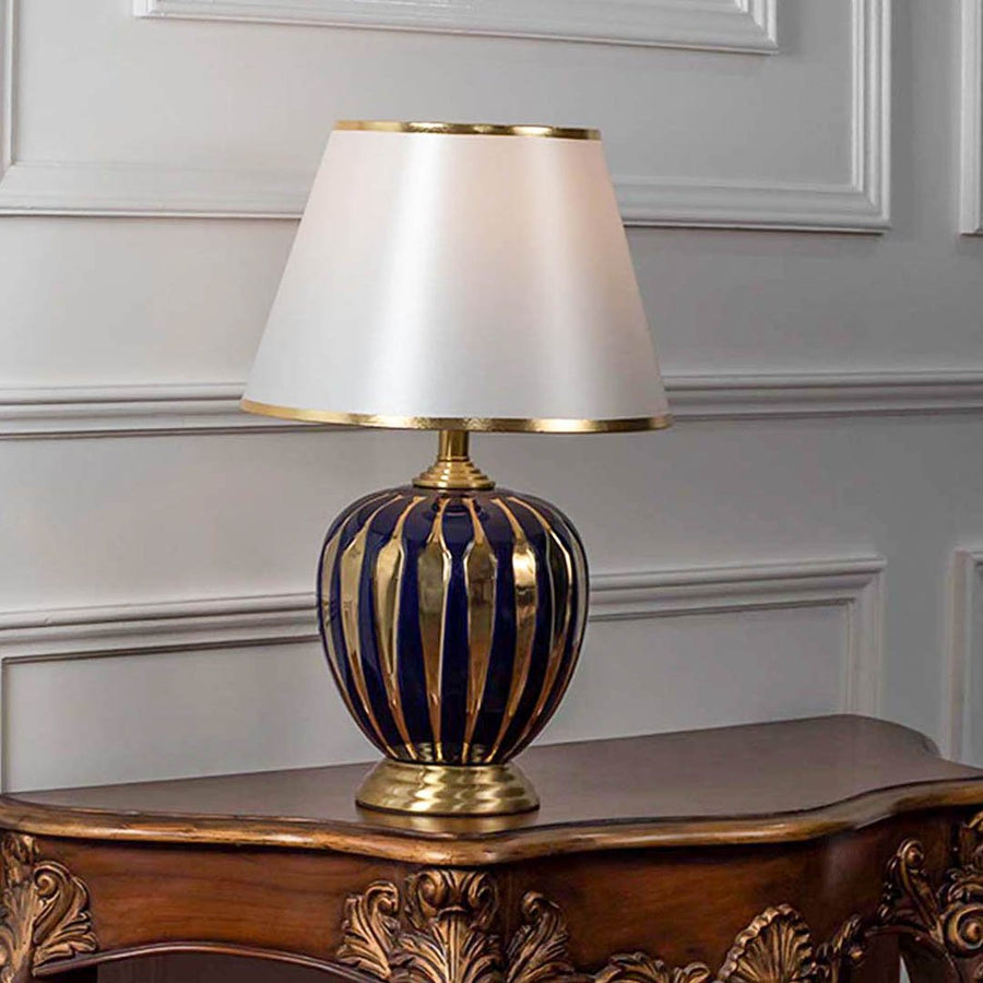 Table lamp with blue glass base and gold accents with white fabric shade on wooden table luxury lighting for living room decor.