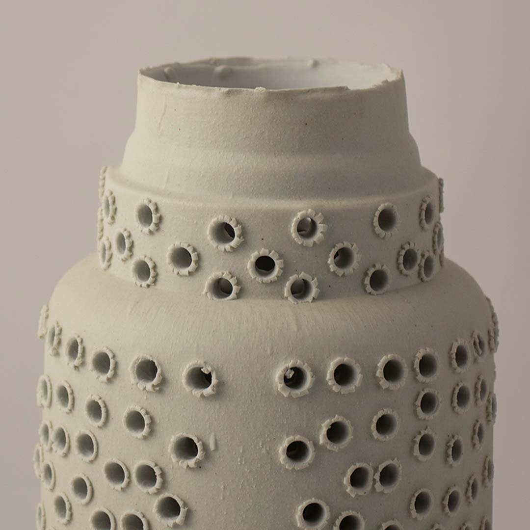 White ceramic lantern vase with perforated hole pattern and lid placed on wooden table against neutral wall background closeup highlighting decorative ambient lighting design.