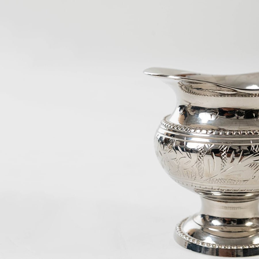 Closeup of silver creamer jug with ornate floral pattern and polished finish shown on a plain white background.
