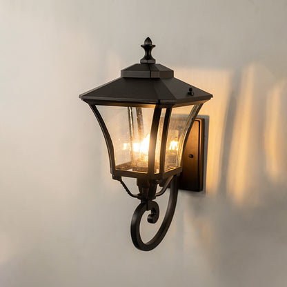 Black metal outdoor wall light on white background featuring a bottom scroll arm and clear glass shade for patio decor.